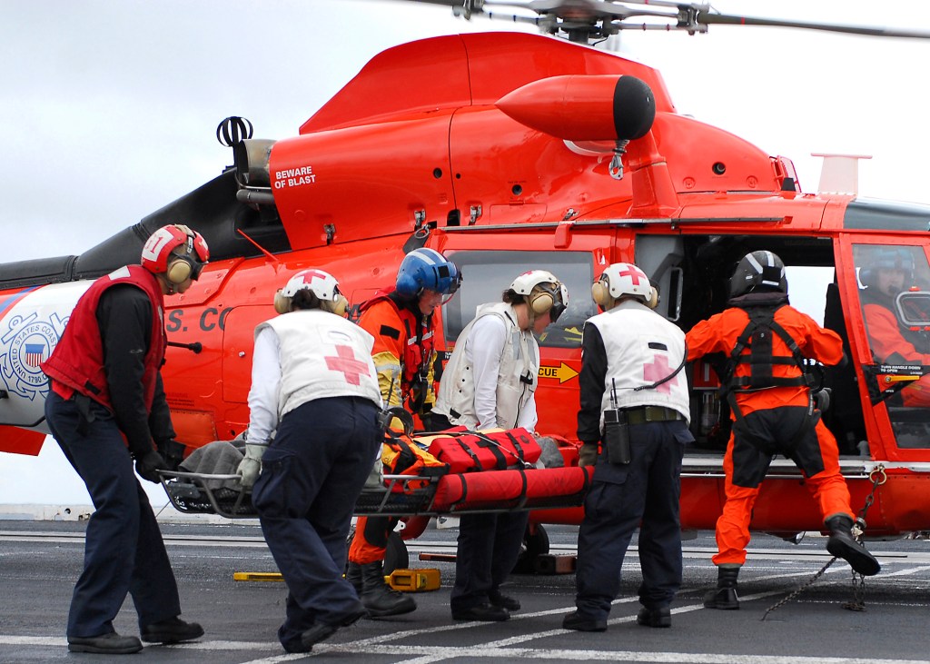 Vier Rettungskräfte transportieren einen Patienten auf einer Trage zum Hubschrauber. Der Hubschrauber ist rot mit einem weißen Emblem und steht auf einer Landefläche. Die Rettungskräfte tragen spezielle Schutzkleidung und Helme.