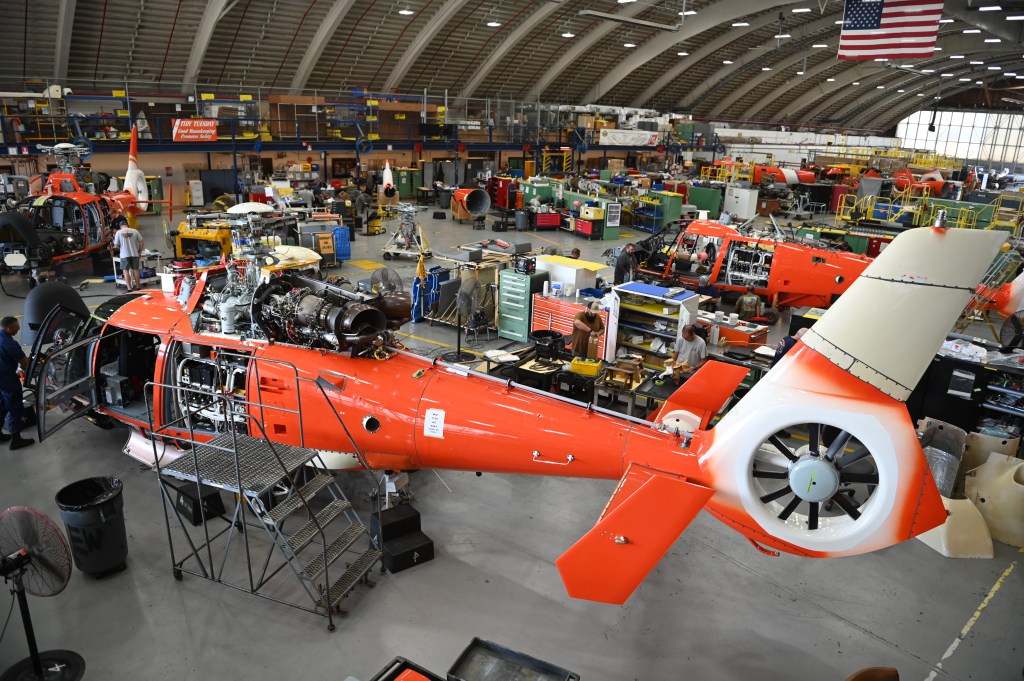 Ein Hubschrauber im Montagewerk mit geöffnetem Cockpit; Arbeiter führen Wartungsarbeiten durch. Im Hintergrund sind weitere orangefarbene Hubschrauber und Werkbänke sichtbar.