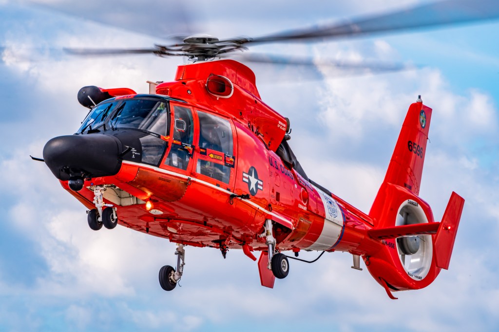 Ein rot-weißer Hubschrauber der US-Küstenwache schwebt in der Luft mit rotierenden Rotorblättern vor einem wolkigen Himmel.