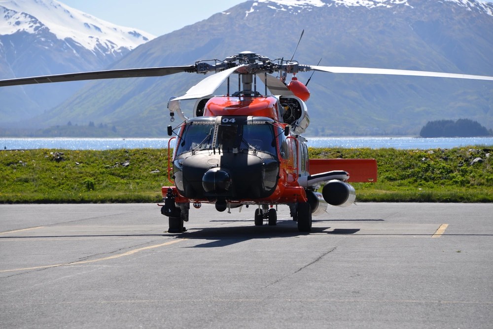 Ein roter und schwarzer Hubschrauber steht auf einem Parkplatz mit schneebedeckten Bergen im Hintergrund und einem Gewässer in der Nähe.