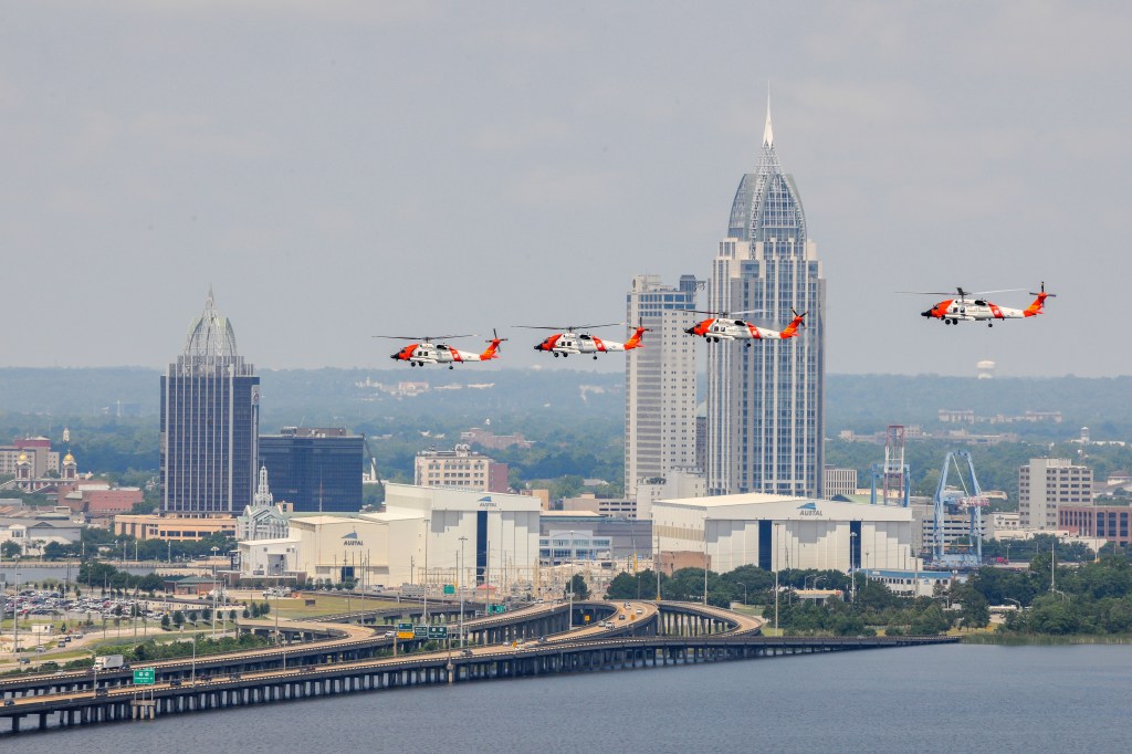 Mehrere Hubschrauber fliegen über die Skyline einer Stadt mit modernen Wolkenkratzern und einer Brücke im Vordergrund.