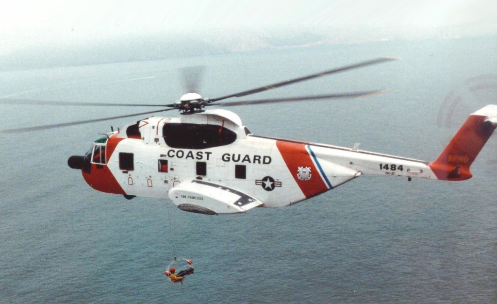 Ein Hubschrauber der Küstenwache fliegt über das Wasser, während ein Rettungseinsatz mit einem Seilrettungspersonal durchgeführt wird.