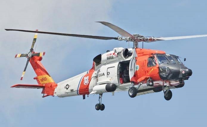 Ein Hubschrauber der Küstenwache fliegt am blauen Himmel, mit orange-weißer Farbgebung und offenen Türen.
